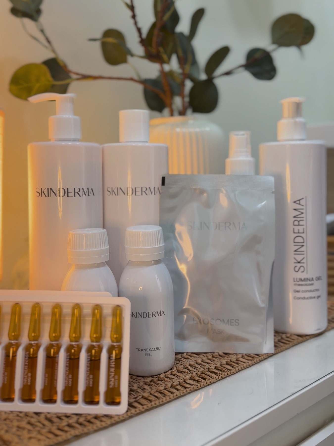 Botes, ampollas y una mascarilla en sobre de cosmética Skinderma sobre una cómoda blanca, con ramas de eucalipto detrás