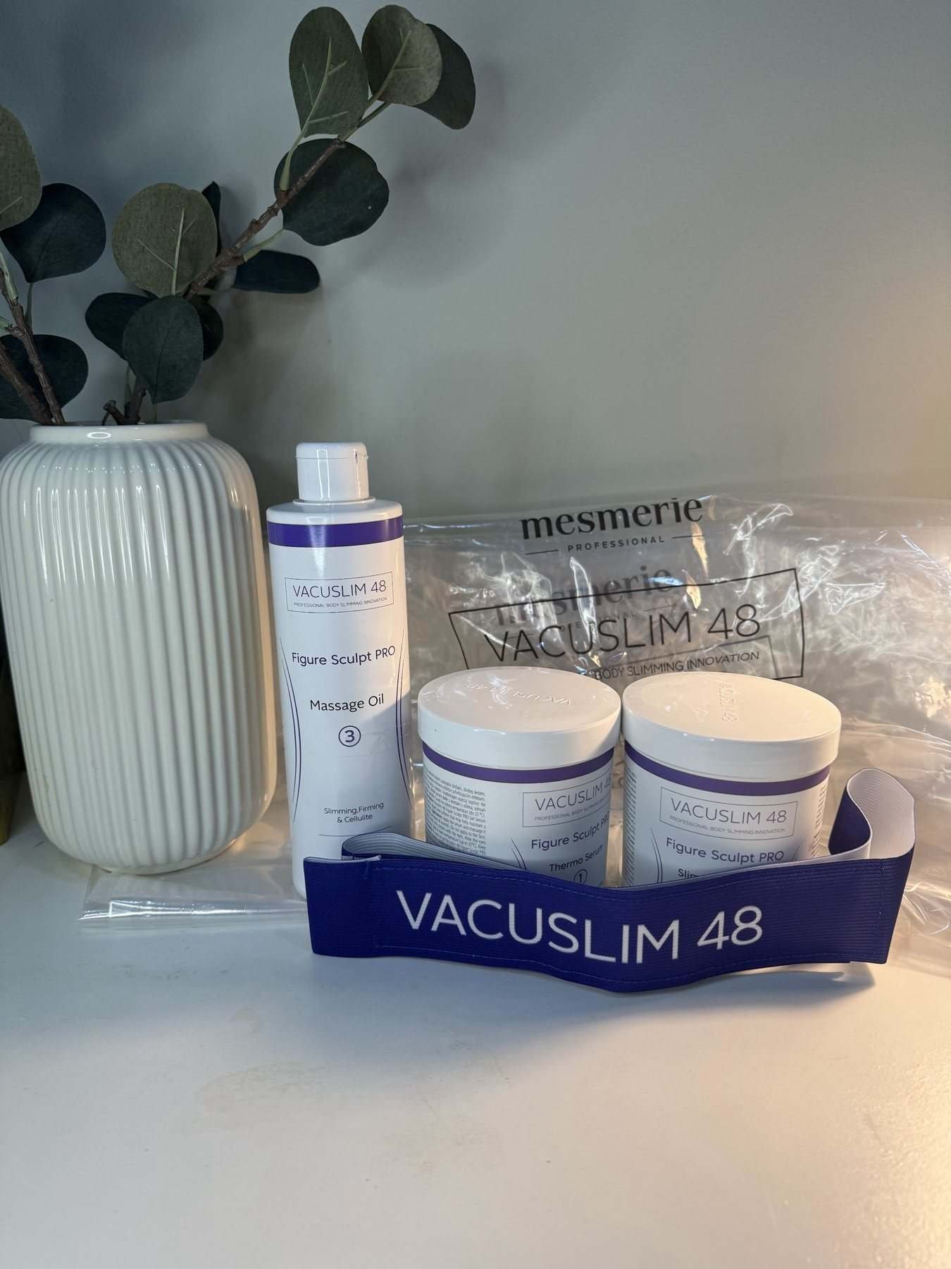 Productos de Vacuslim 48 sobre una mesa blanca, junto a un jarrón con ramas de eucalipto
